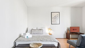 Ein modernes Hotelzimmer mit einem ordentlich bezogenem Bett, einem Nachttisch mit Lampe, einem Sessel, einem Schreibtisch mit Computer und einem gerahmten Kunstwerk an der Wand.