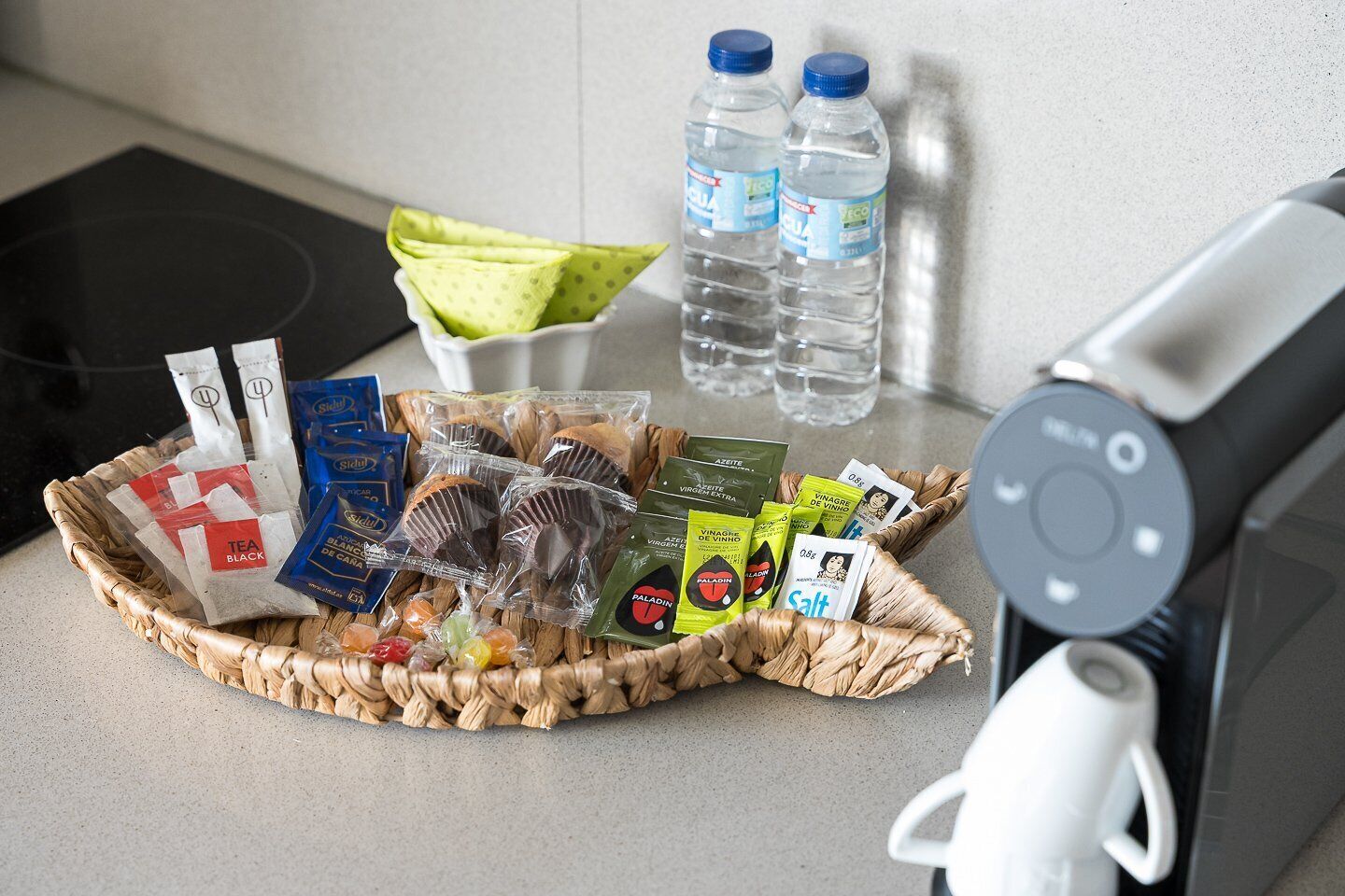 Un piano di lavoro in cucina con un cesto contenente tè in busta, cioccolatini, snack e bottiglie d'acqua.