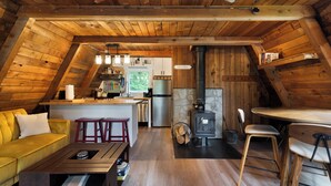 A cozy attic-style living space with a wooden ceiling, a sofa, a dining table, and a kitchen area with a stove.