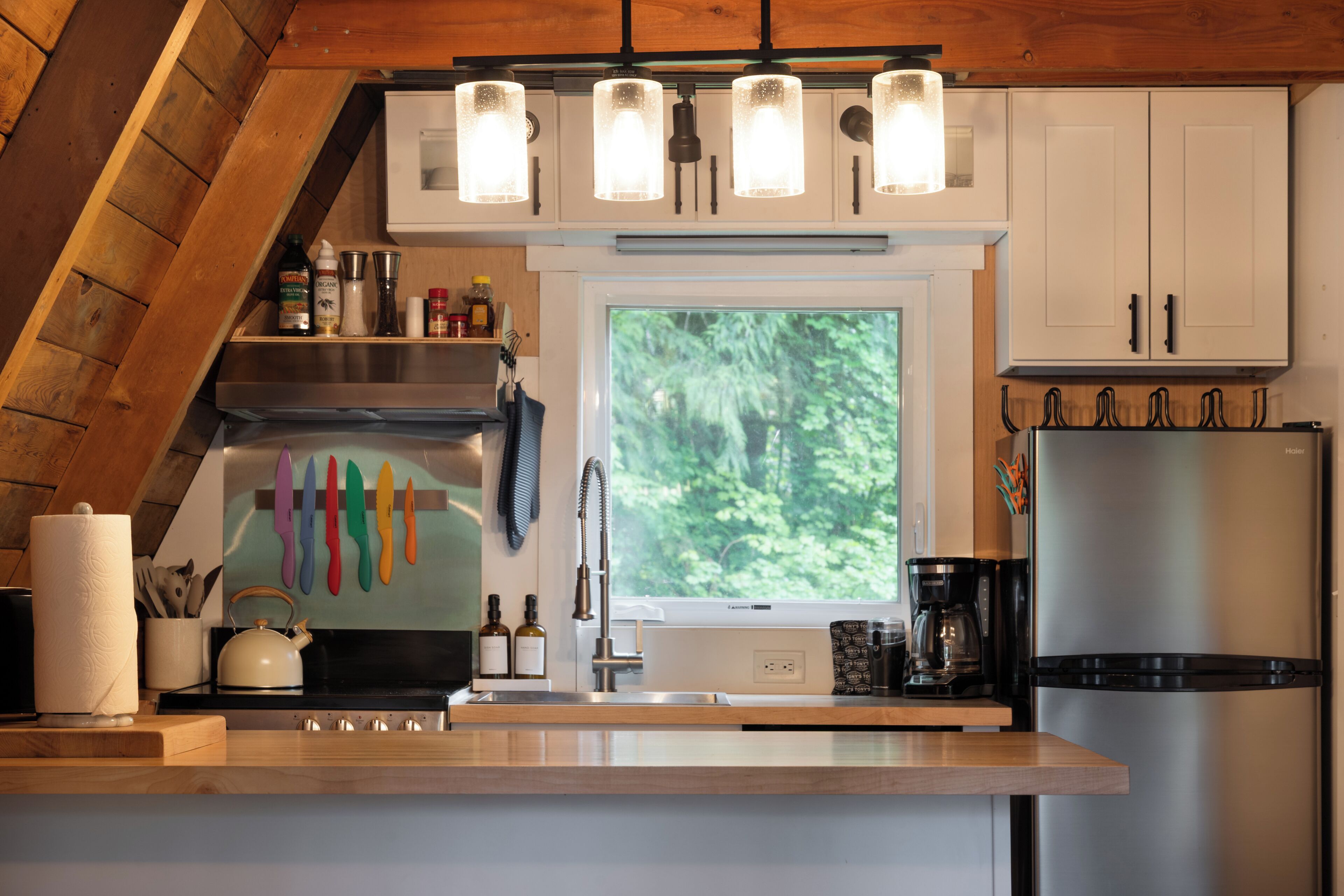 A modern kitchen with a wooden ceiling, white cabinets, a stainless steel refrigerator, and a window with a view of greenery.