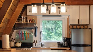 A modern kitchen with a wooden ceiling, white cabinets, a stainless steel refrigerator, and a window with a view of greenery.