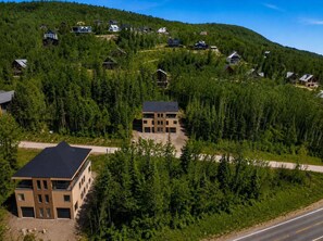 A residential area with multiple houses surrounded by dense forest, a road, and a modern two-story building with a flat roof. - Mountain | Sauna | Camp de base du Massif (Saint-Philémon)
