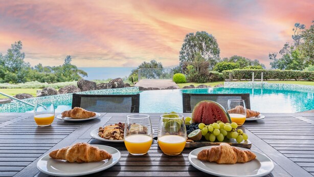 Una mesa de desayuno con croissants, fruta y jugo, con vista a una piscina y un paisaje pintoresco.