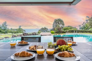 Ett frukostbord med croissanter, frukt och juice, med utsikt över en pool och ett vackert landskap.