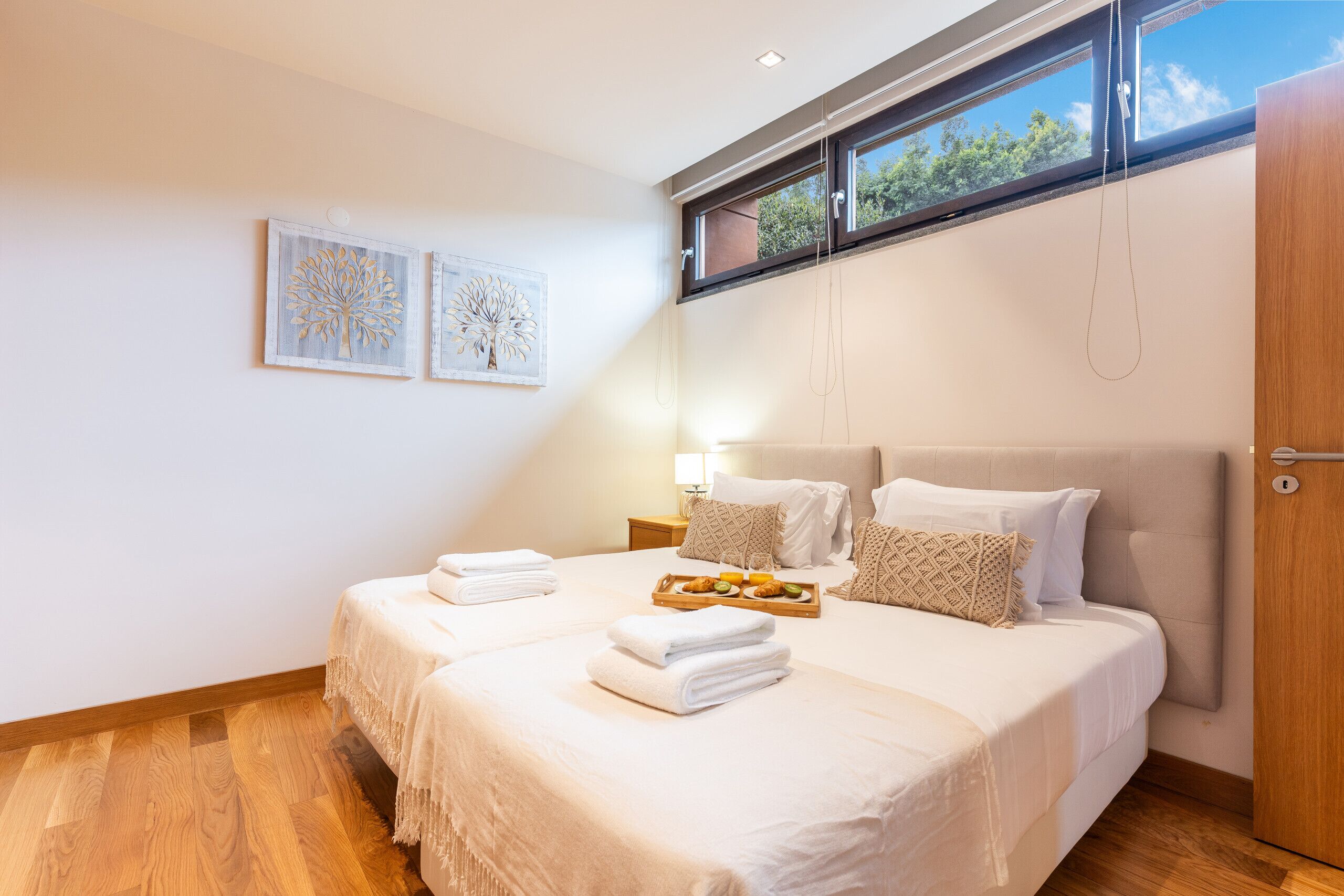 A modern hotel room with two beds, a wooden floor, and a large window with a view of trees.
