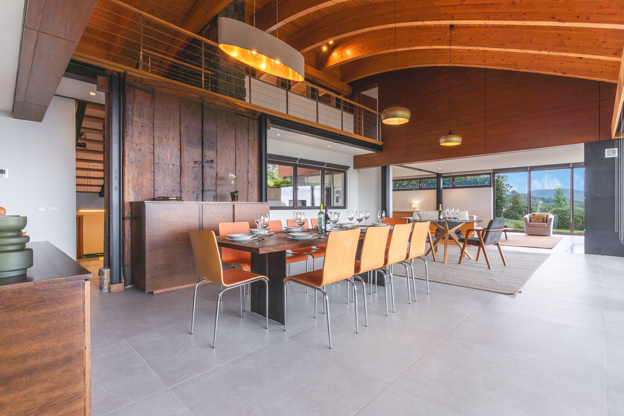 Un espace de salle à manger moderne, avec une longue table en bois, de nombreuses chaises et un plafond haut avec des poutres apparentes.