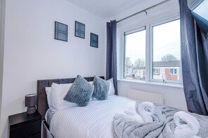 Un dormitorio con cama, almohadas, mesita de noche con lámpara, ventana con cortinas y vistas a edificios.