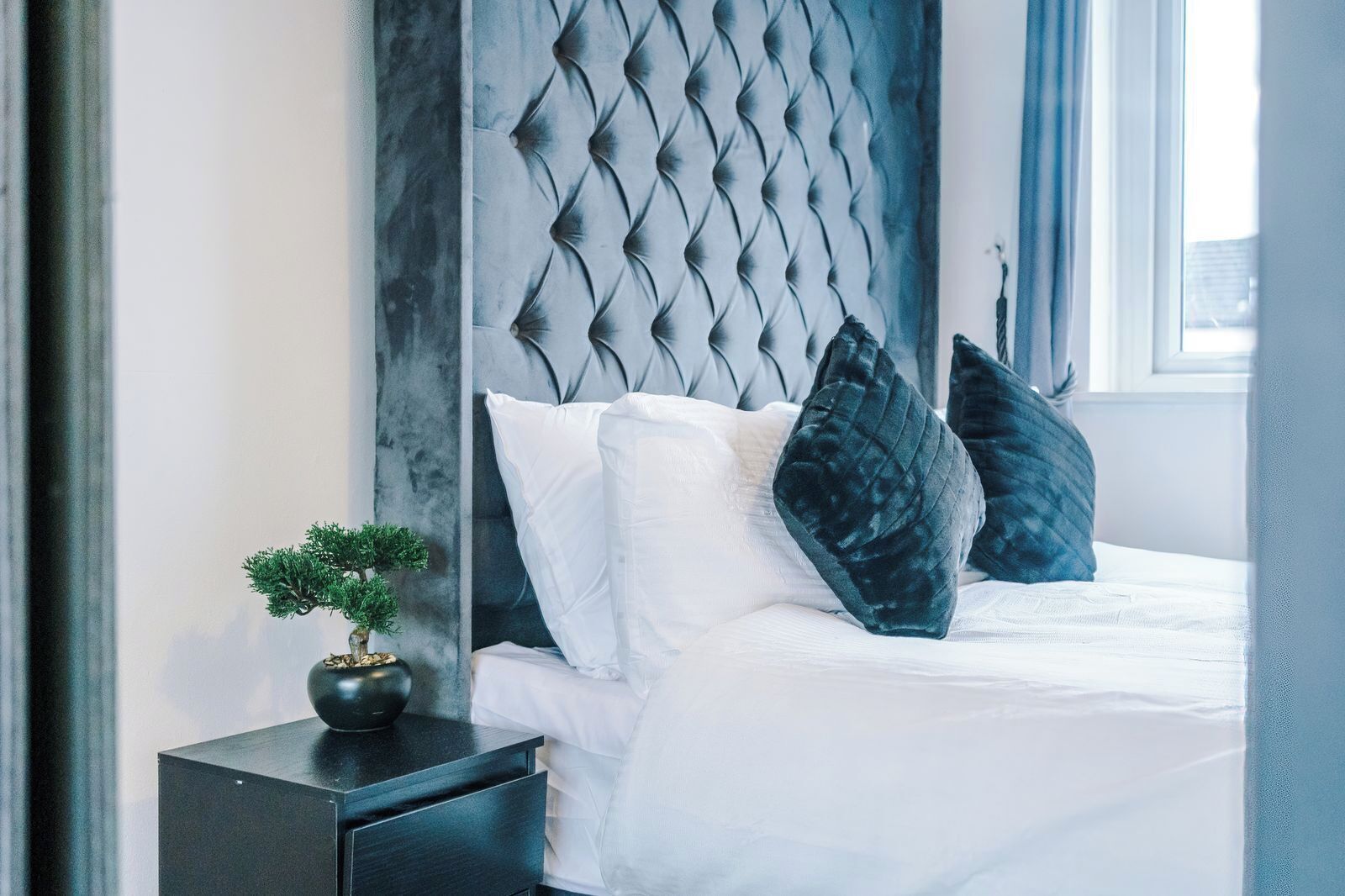 A neatly made bed with white bedding and dark pillows, a small bedside table with a plant, and a dark headboard.