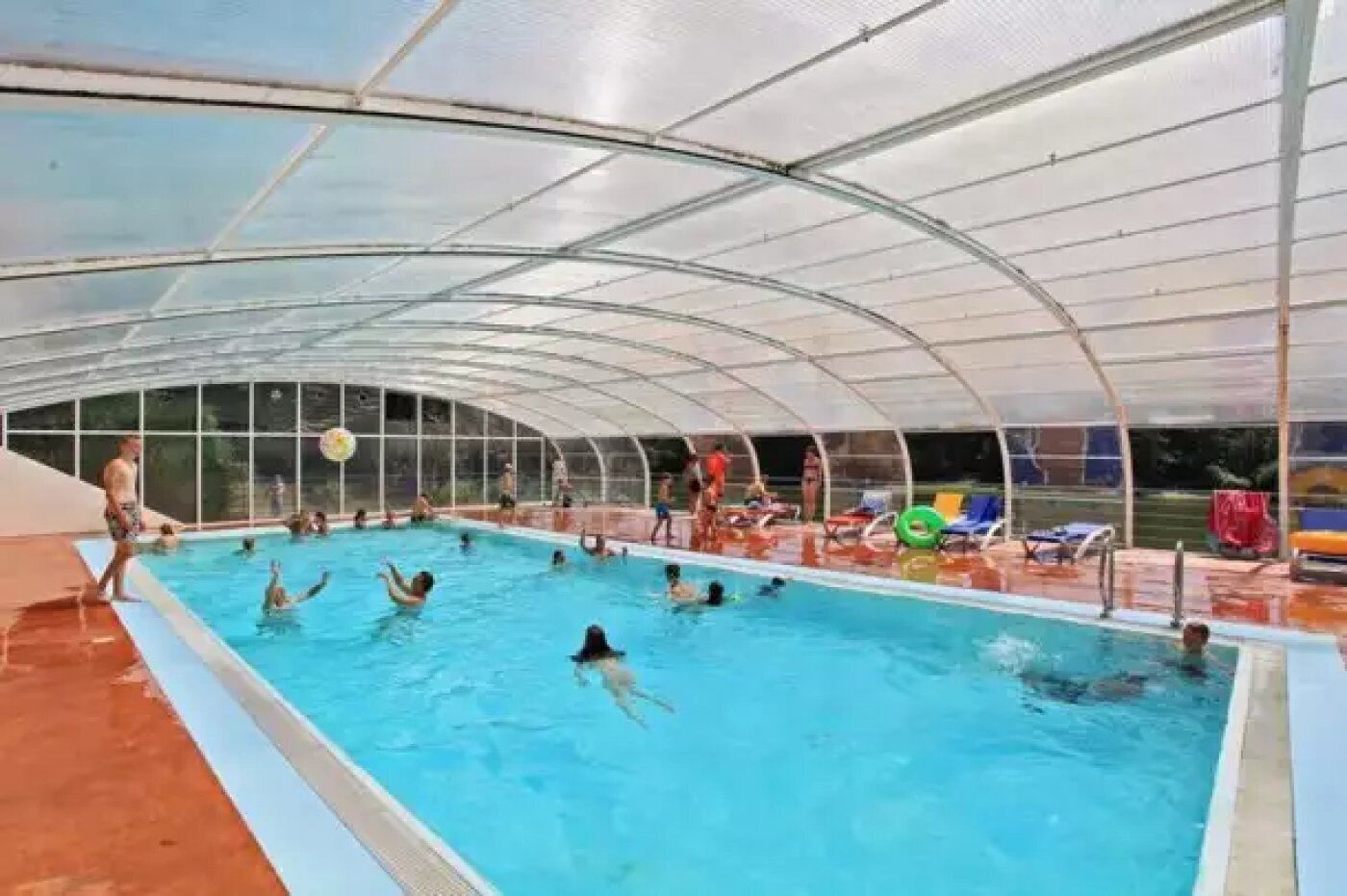An indoor swimming pool with a transparent roof, surrounded by lounge chairs and a tiled floor.