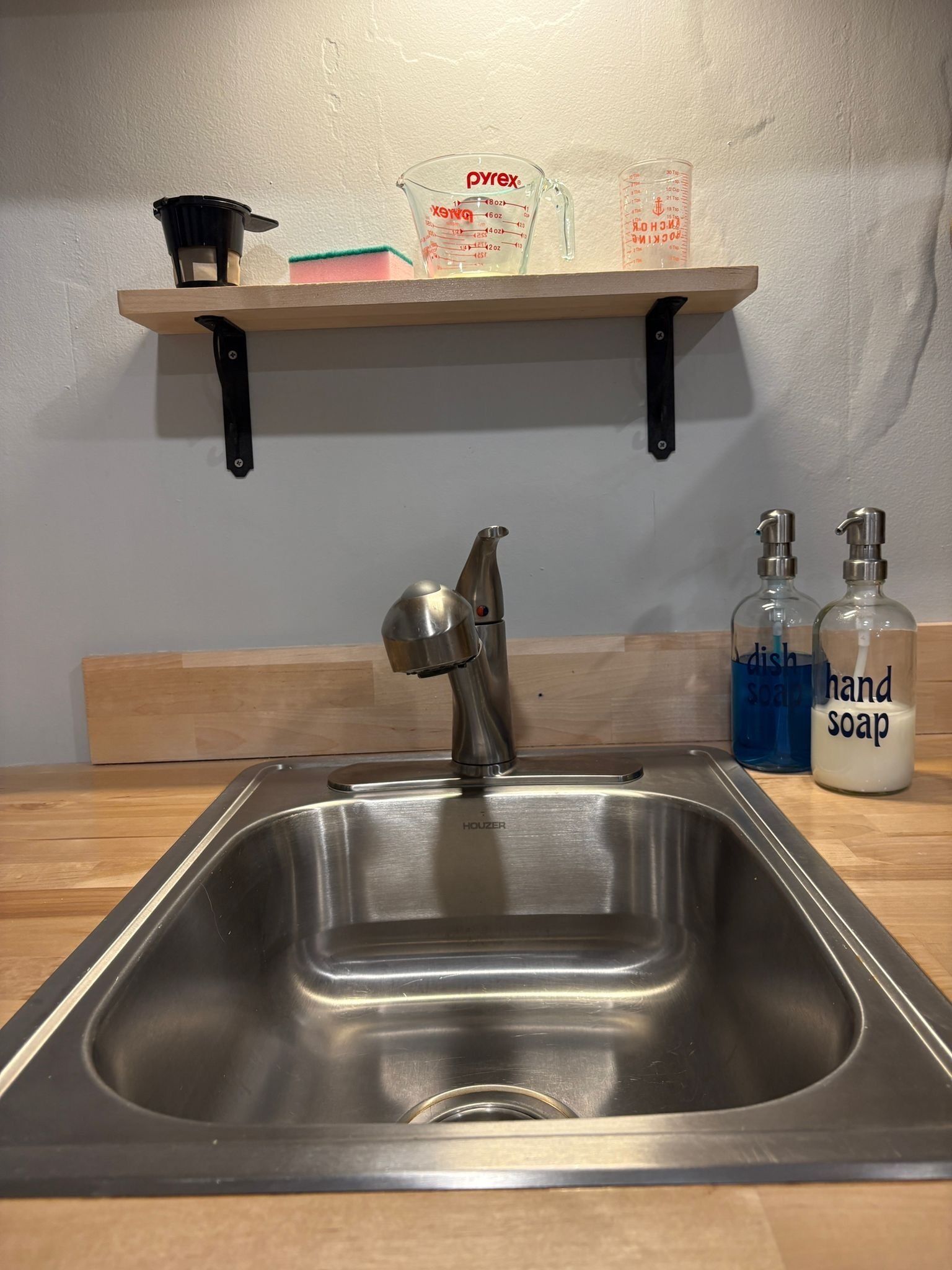 Un lavamanos de cocina con un grifo moderno, un estante de madera arriba con una cafetera, un vaso medidor y una caja de Pyrex.
