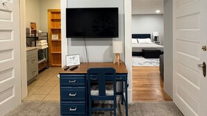 Une pièce comprenant un bureau, une chaise et un téléviseur fixé au mur. La pièce adjacente contient un lit, une table de chevet et une lampe.