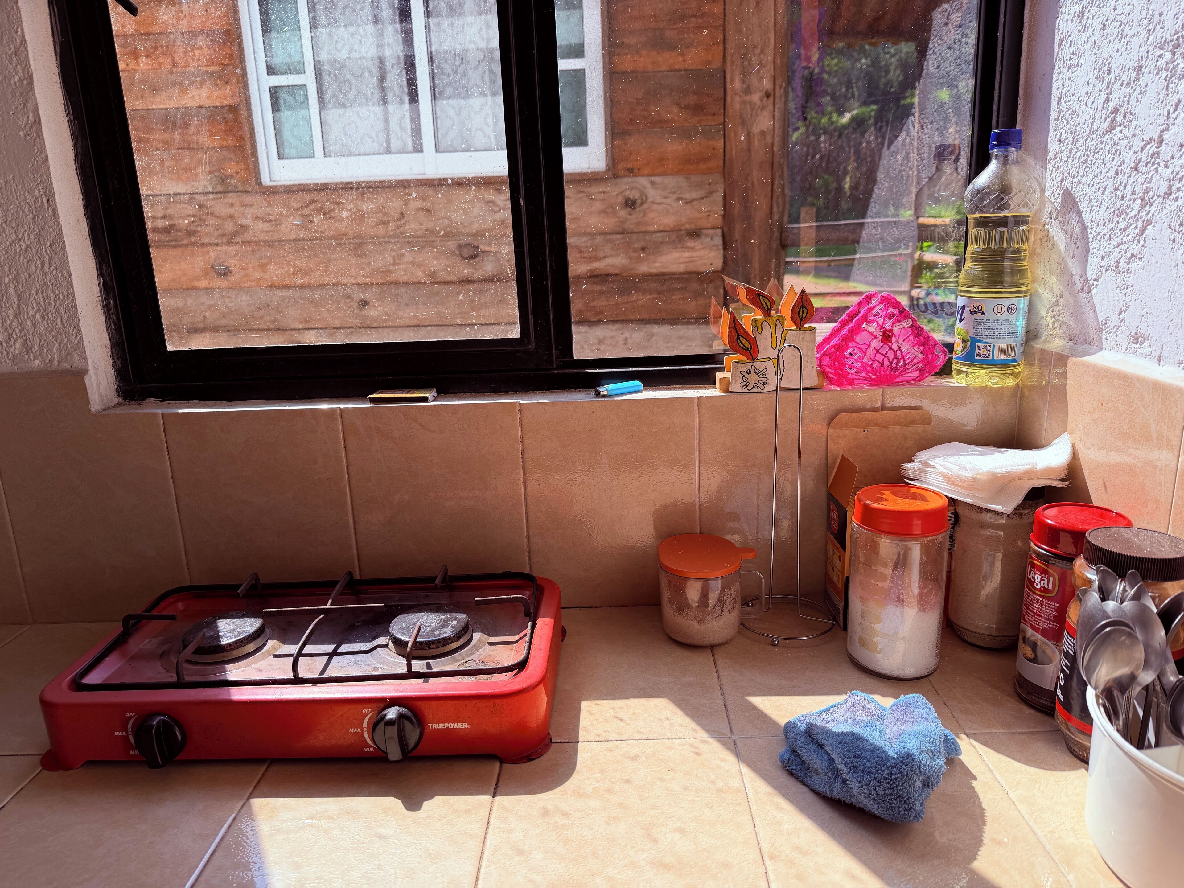 A kitchen with a red gas stove, a window with a view of a wooden cabin, and various kitchen items like a bottle of cooking oil, a jar of sugar, and a blue cloth on the counter.