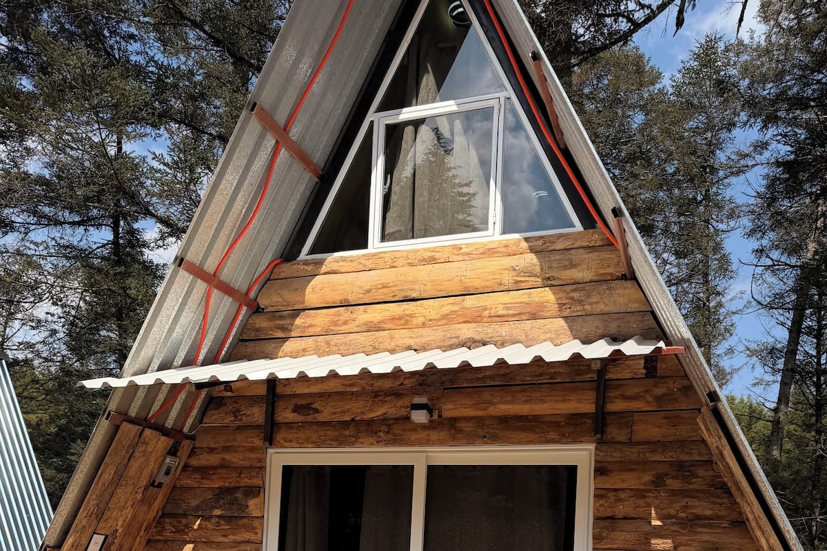 Una cabaña de madera con un amplio ventanal corredera, un pequeño porche con plantas en macetas y un techo a dos aguas.