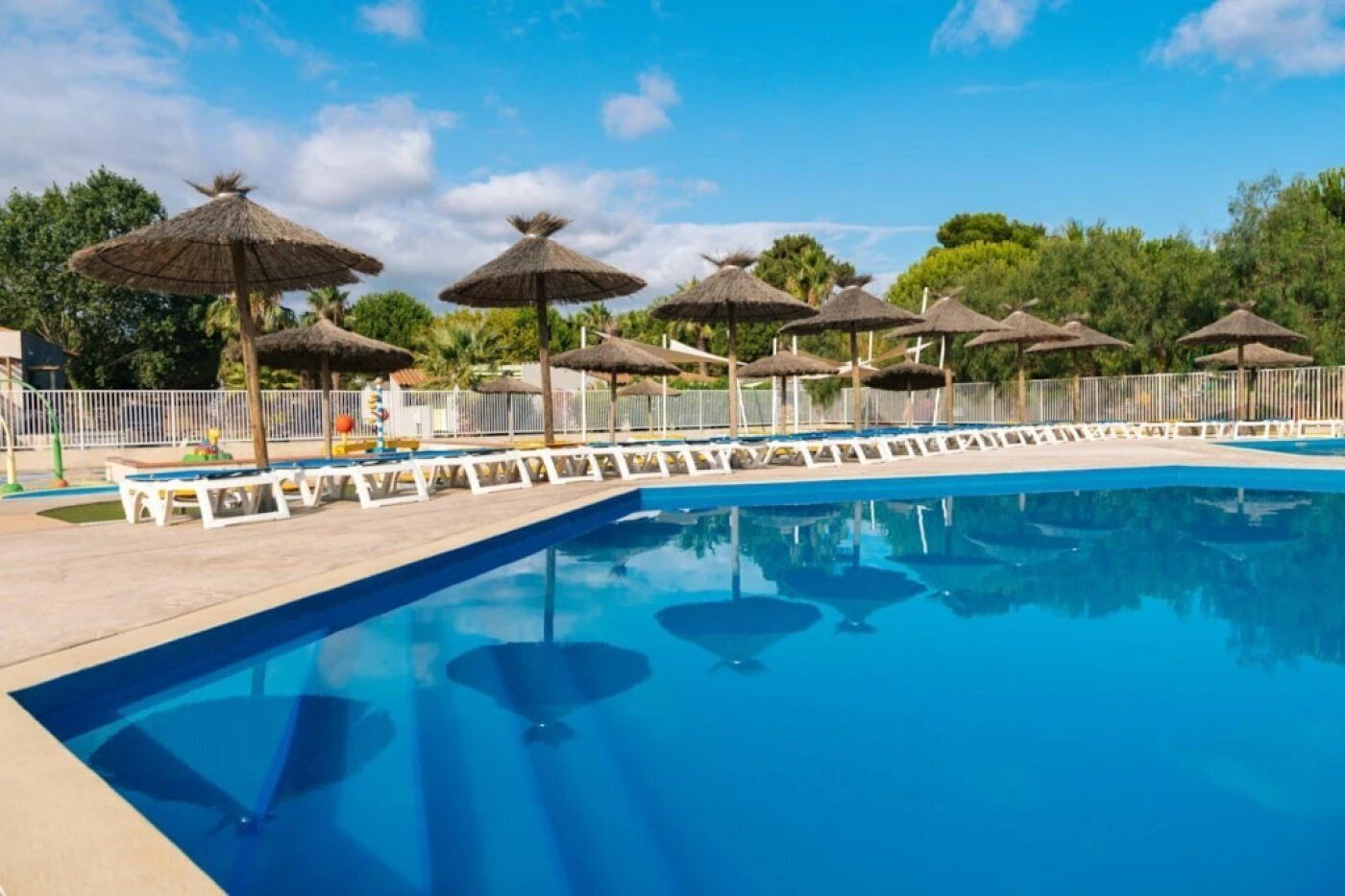 A swimming pool with lounge chairs and thatched umbrellas, surrounded by a fence and greenery.
