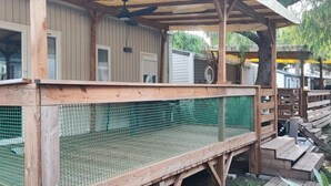 Un'area relax con terrazza in legno, recinzione a rete, ringhiere in legno e un tetto che protegge da sole e pioggia, con una ventola a soffitto.
