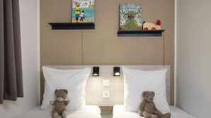 Quarto de hotel com duas camas, cada uma com um travesseiro branco e um ursinho de pelúcia. Existem duas prateleiras fixadas na parede acima das camas, uma com um quadro e outra com um livro e um brinquedo.