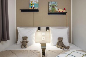 A hotel room with two beds, each with a white pillow and a teddy bear. There are two wall-mounted shelves above the beds, one with a painting and the other with a book and a toy. - ZEcamping 4 étoiles piscine chauffée animations bien-être locatif 29m2 (Saint-Jean-de-Monts)
