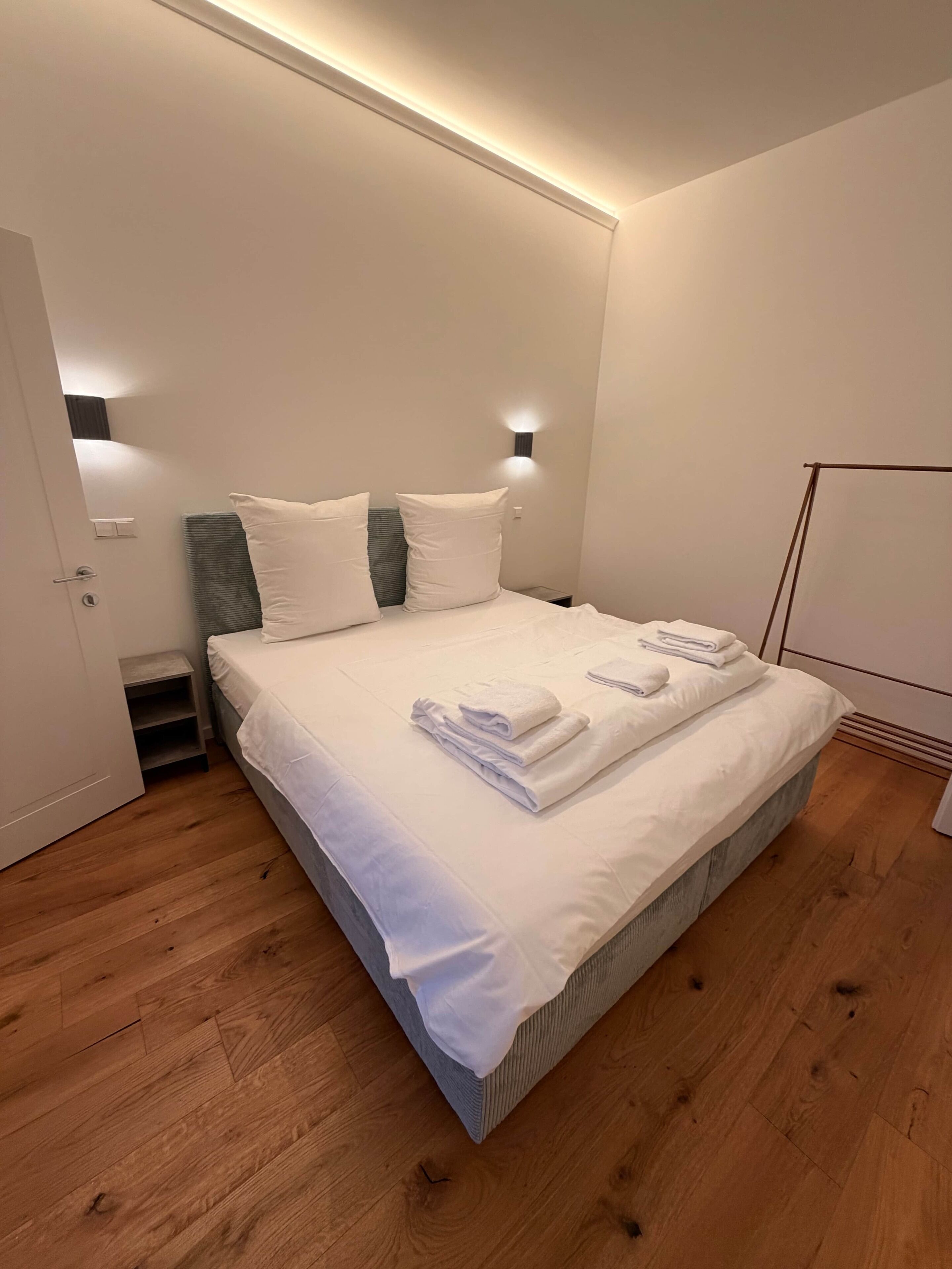 Ein ordentlich bezogenes Bett mit weißen Leinen und Handtüchern, ein Nachttisch und eine Wandleuchte.