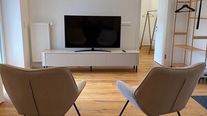 Ein modernes Wohnzimmer mit Flachbildfernseher, zwei Sesseln, einer weißen Unterhaltungsecke und Holzboden.