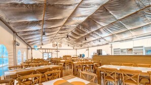 Een ruime, overdekte eetruimte met houten tafels en stoelen, een hoge plafond met metalen constructie, en grote ramen die natuurlijk licht binnenlaten.