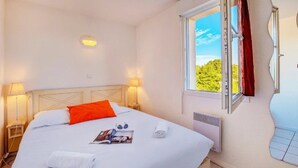 Een keurig opgemaakt bed met witte lakens en een rood kussen, een bedlampje, een raam met uitzicht op groen en een wandradiator.