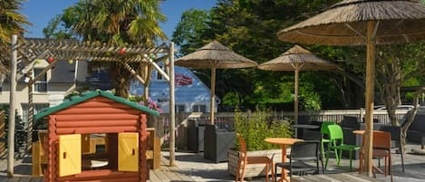 A wooden deck with a small playhouse, outdoor seating, and thatched umbrellas.