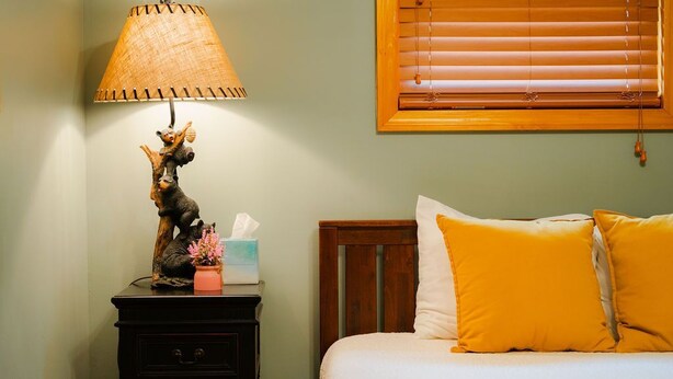 Een slaapkamer met een bed, een nachtkastje met een lamp en een raam met jaloezieën.