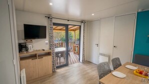 Una cucina moderna con TV, macchina del caffè e mobili in legno. C'è un'area pranzo con tavolo, sedie e una ciotola di frutta. Una porta scorrevole in vetro dà accesso a un terrazzo esterno con tavolo e sedie.