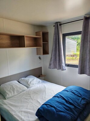 Un lit simple avec une couette blanche et bleue, une tête de lit en bois et une petite table de chevet. Il y a des étagères en bois au-dessus du lit et une fenêtre avec des rideaux.