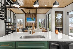 Une cuisine moderne dotée d’un grand îlot central, d’une cuisinière intégrée et d’un évier. Un escalier en colimaçon se trouve à gauche et une grande fenêtre à droite.