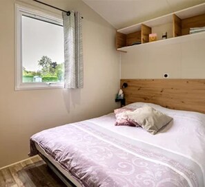 A bedroom with a bed, a window with curtains, a wooden headboard, and a shelf above the bed. - ZEcamping 3* mobil-home TV BBQ 27m2 4 pers (Pordic)