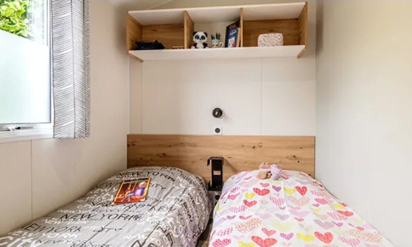 A bedroom with two beds, a wooden headboard, a shelf above the beds, and a window on the left.
