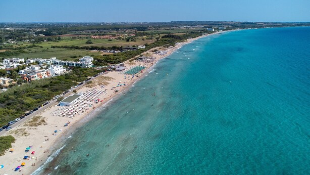 Una vista aerea di una zona costiera con spiaggia, edifici e acqua cristallina blu.