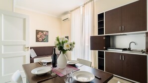 Un'area pranzo con un tavolo apparecchiato per due persone, un vaso con fiori, una bottiglia e un quadro appeso al muro.