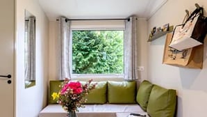 A cozy room with a window, a sofa with green cushions, a small round table with a vase of flowers, and a shelf with various items. - ZEcamping 3 étoiles mobil-home TV LV BBQ 27m2 4 pers (Pordic)