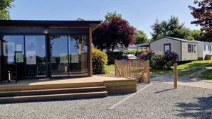 A modern building with a glass entrance, a wooden deck, and a gravel path leading to a fenced area with a tree and parked cars. - ZEcamping 3 étoiles mobil-home TV LV BBQ 27m2 4 pers (Pordic)