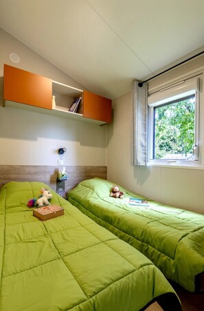 A bedroom with two beds, a shelf with books, a window with curtains, and a small table with a lamp. - ZEcamping 3* chiens acceptés jacuzzi mobil-home TV BBQ 27m2 4 pers (Pordic)