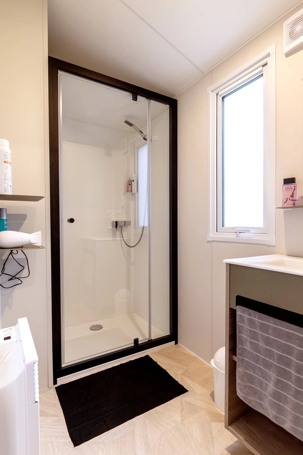 Une salle de bain moderne avec une douche à parois de verre, un lavabo blanc et une fenêtre laissant entrer la lumière naturelle.