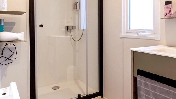A modern bathroom with a glass-enclosed shower, a white sink, and a window providing natural light. - ZEcamping 3* chiens acceptés jacuzzi mobil-home TV BBQ 27m2 4 pers (Pordic)