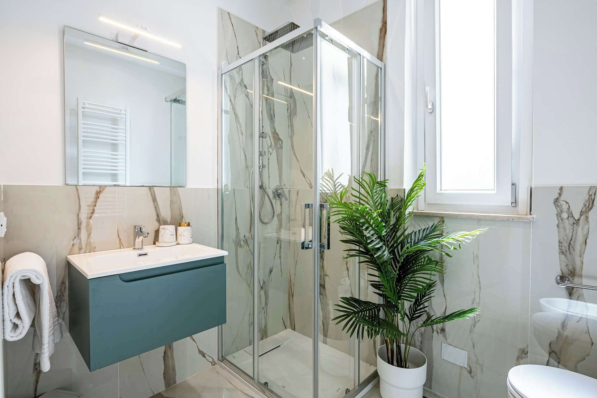 Un baño moderno con cabina de ducha de vidrio, pared con azulejos de mármol, lavabo con mueble azul y una planta en maceta.