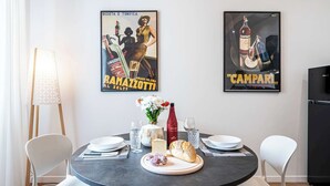 A dining area with a round table set for two, featuring a bottle of red wine, a loaf of bread, and a plate of cheese. Two posters adorn the wall, one promoting 'BAMAZZOTTI' and the other 'CAMPARI'. - Casa Belfiore in Firenze (Florence)