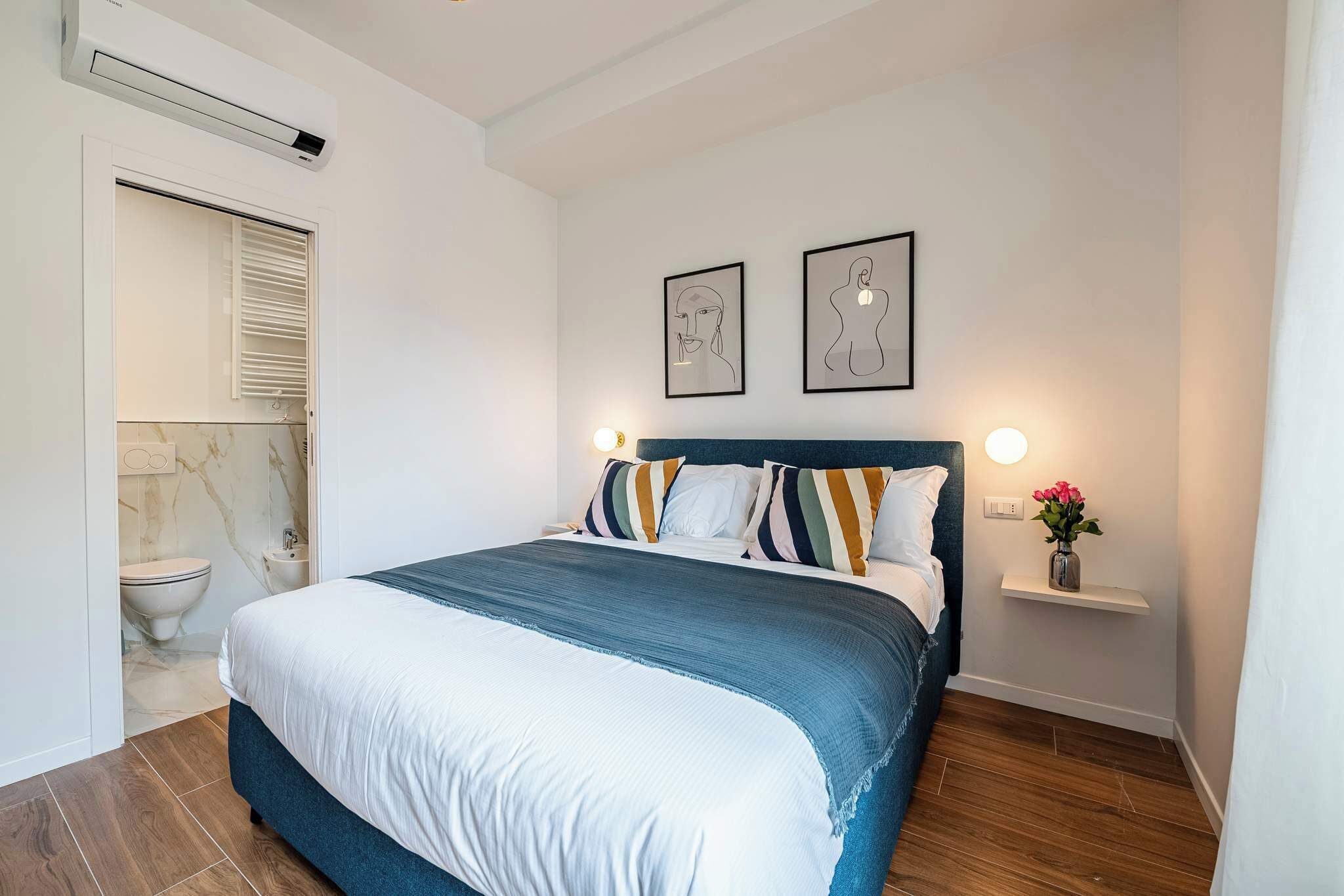 Une chambre à coucher avec un lit, une table de chevet sur laquelle se trouve un vase, et deux tableaux encadrés accrochés au mur.