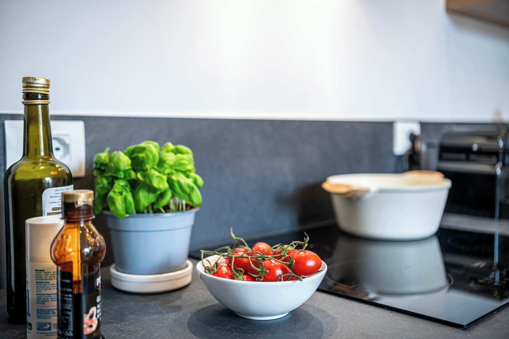 Eine Küchenarbeitsplatte mit einer Schüssel voll Kirschtomaten, einer Olivenölflasche, einer kleinen Saucenflasche und einem Topf mit Basilikum.