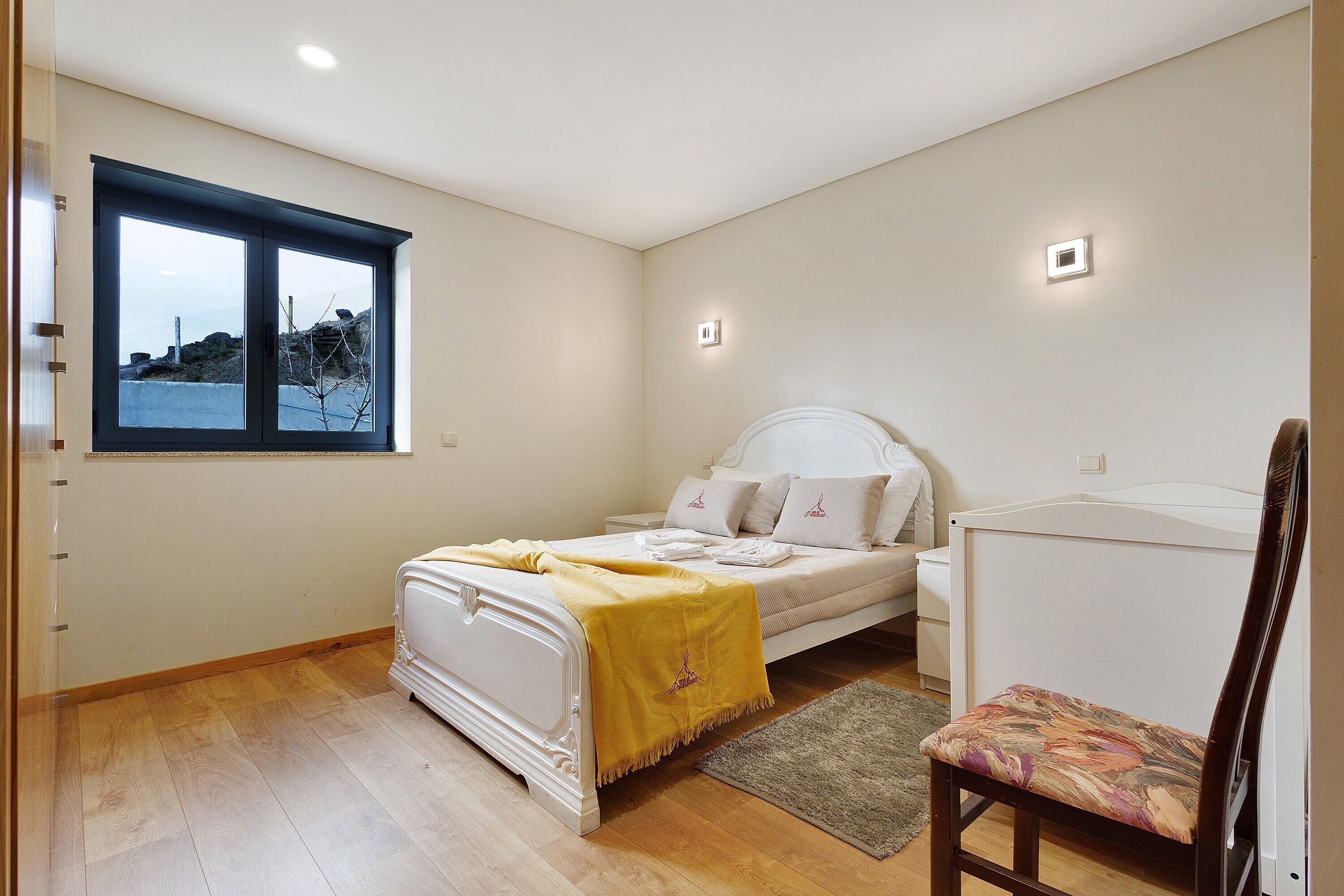 Ein Schlafzimmer mit einem Bett, einem Sessel, einem Fenster und einem kleinen Nachttisch.