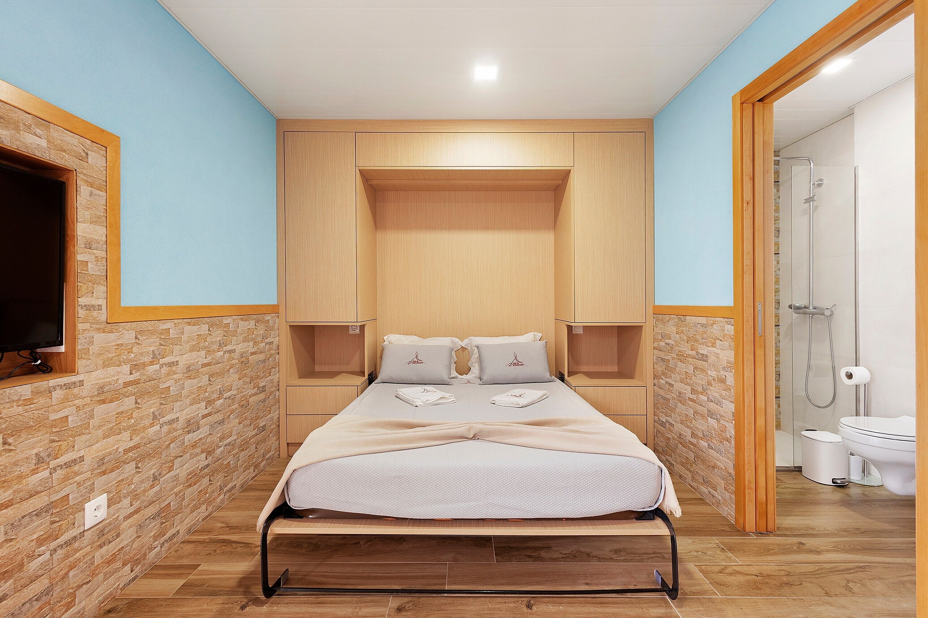 Ein Schlafzimmer mit Bett, Holz-Kopfstück, Nachttischen, einem Fernseher und einem Badezimmer mit Dusche und Toilette.