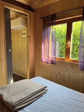 A wooden room with a bed, a window with curtains, and a door leading to another room.