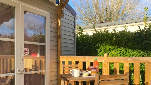 A wooden deck with a table and chairs, a covered area with a roof, and a view of a hedge and trees in the background. - ZEcamping 2* chiens acceptés locatif TV clim 20m2 2 pers (Angicourt)