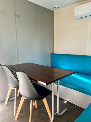 A modern room with a brown table, two grey chairs, a blue bench, and a white air conditioning unit. - ZEcamping 2 étoiles chiens acceptés locatif TV clim 20m2 2 pers (Angicourt)