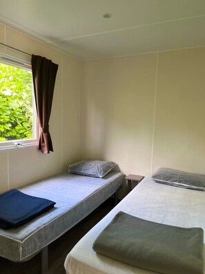 A dormitory room with two single beds, a window with curtains, and a view of greenery outside. - ZEcamping 2 étoiles chiens acceptés locatif TV clim 20m2 2 pers (Angicourt)