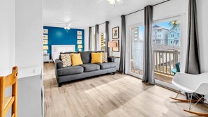 En moderne stue med en grå sofa, trægulv, ventilator og en balkon med udsigt til huse.